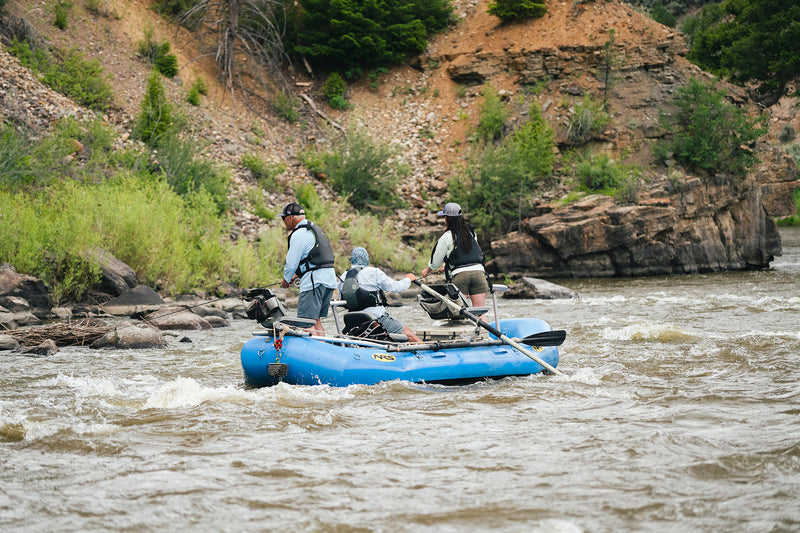 Three people in a blue raft navigating a river while fishing with rocky banks and greenery.