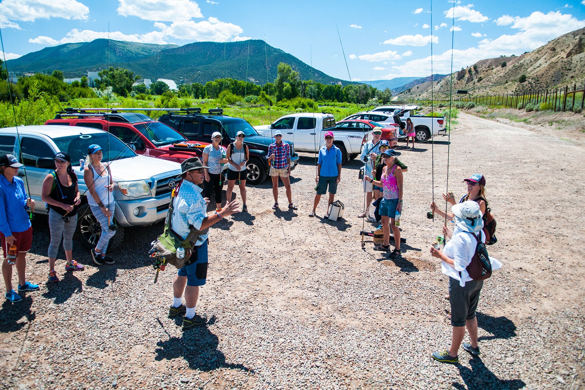 A group of students with fly fishing gear in a parking lot, with a Vail Valley Anglers instructor with mountains in the background.