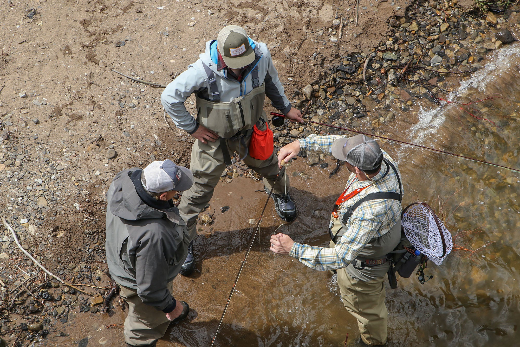 Three people fishing by a stream, with one person demonstrating or explaining technique to the others.