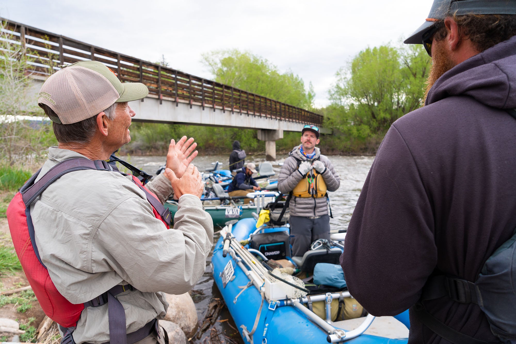 Two men preparing for a river rafting trip with a guide, near a bridge and trees.