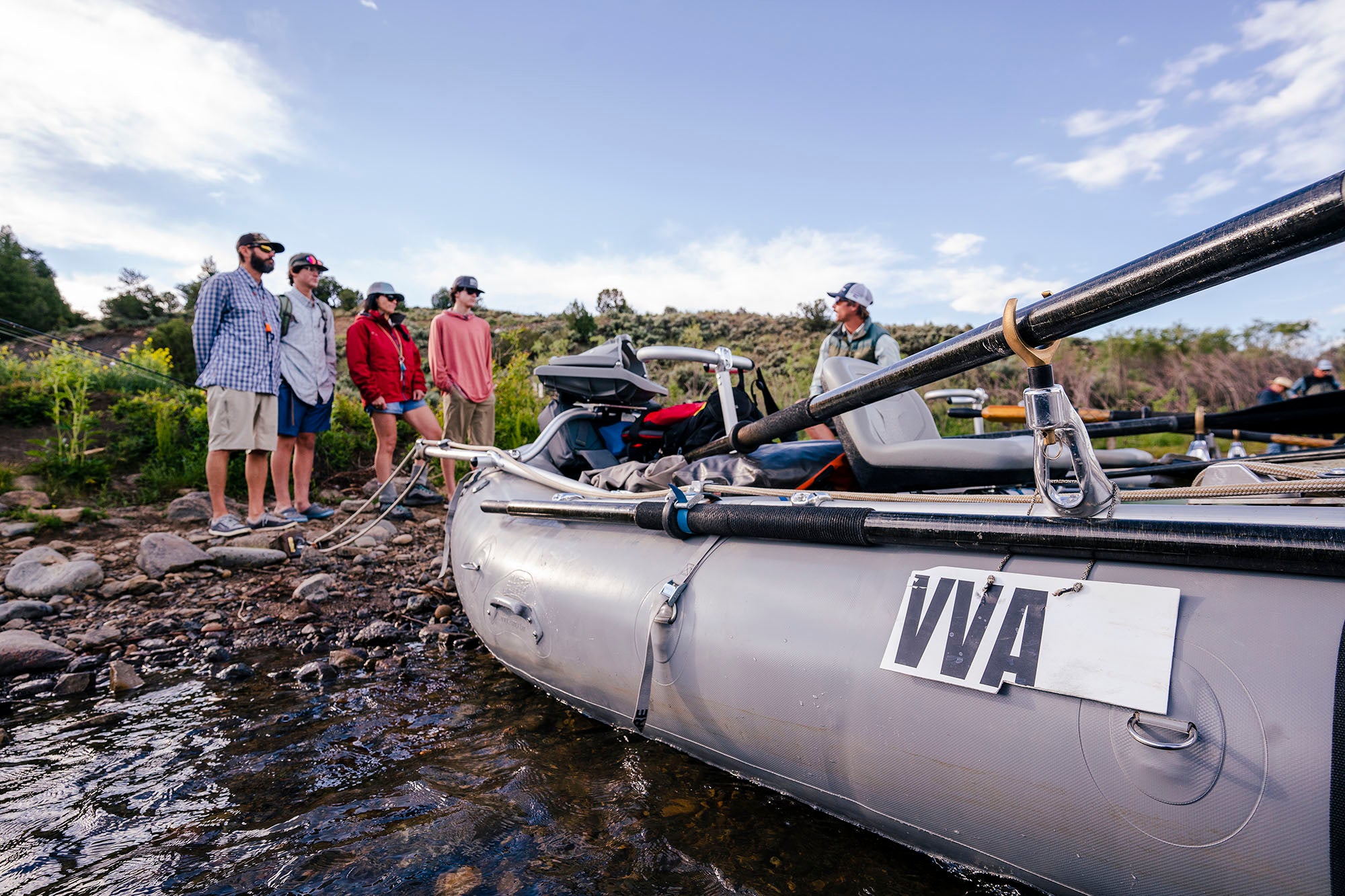 Group of people preparing to go fly fishing with a Vail Valley Anglers raft on a riverbank.