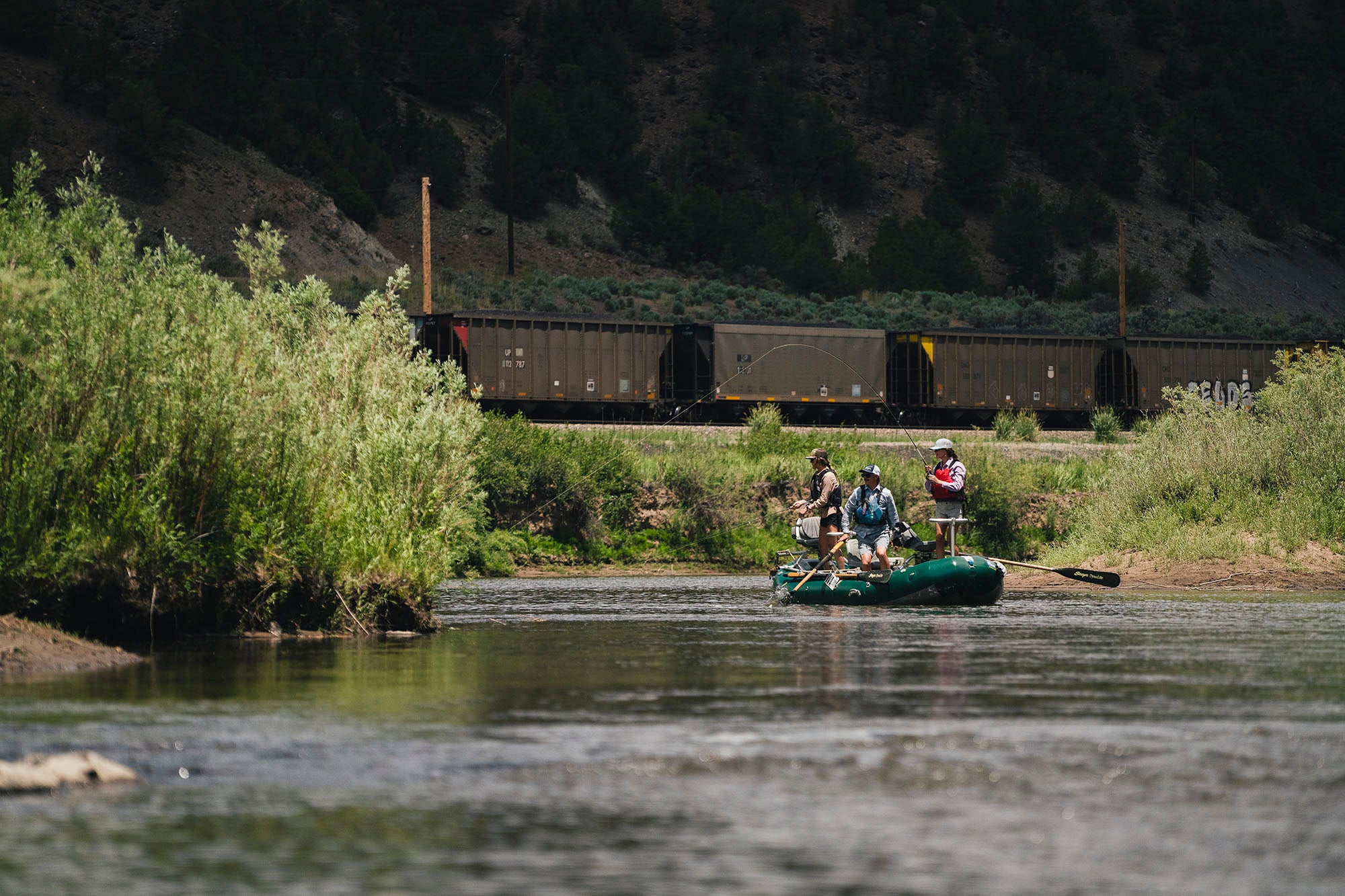 Three people in a raft on a river with a train in the background