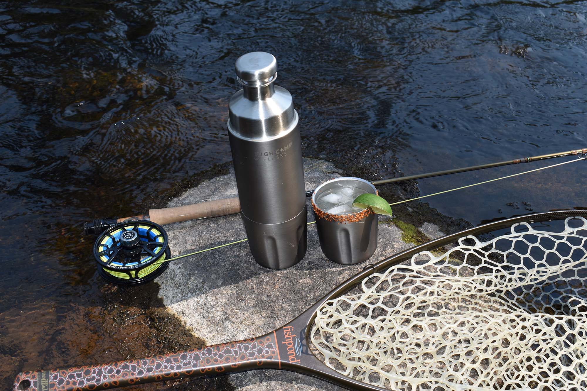 A fly fishing setup on a rock in flowing water, featuring a flask from High Camp Flasks, a Hardy  fly rod and reel, and a Fishpond landing net in the foreground.