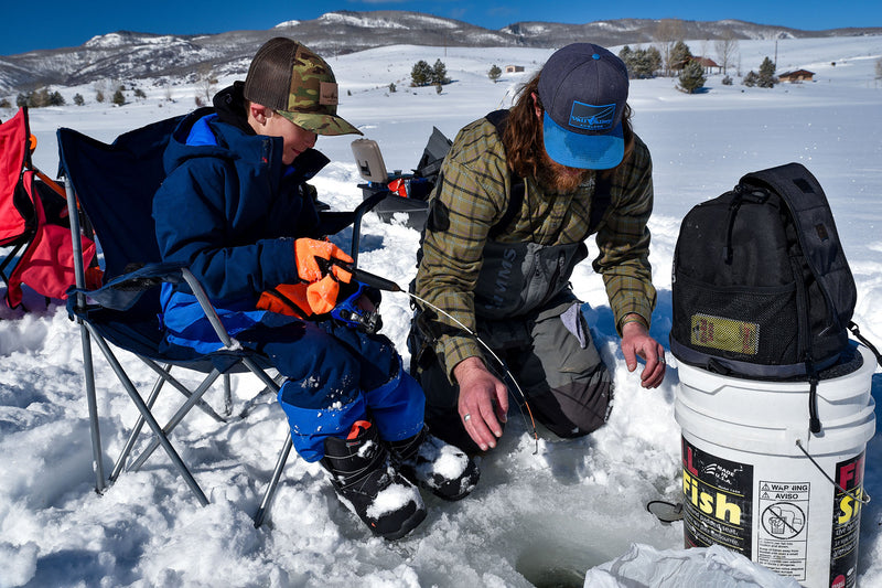Two people ice fishing in a snowy landscape with mountains in the background.