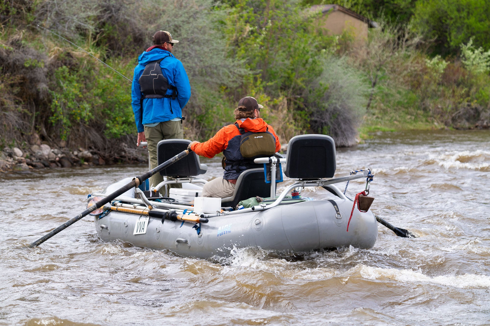 Two people in a small inflatable boat on a river with trees and rocks in the background.