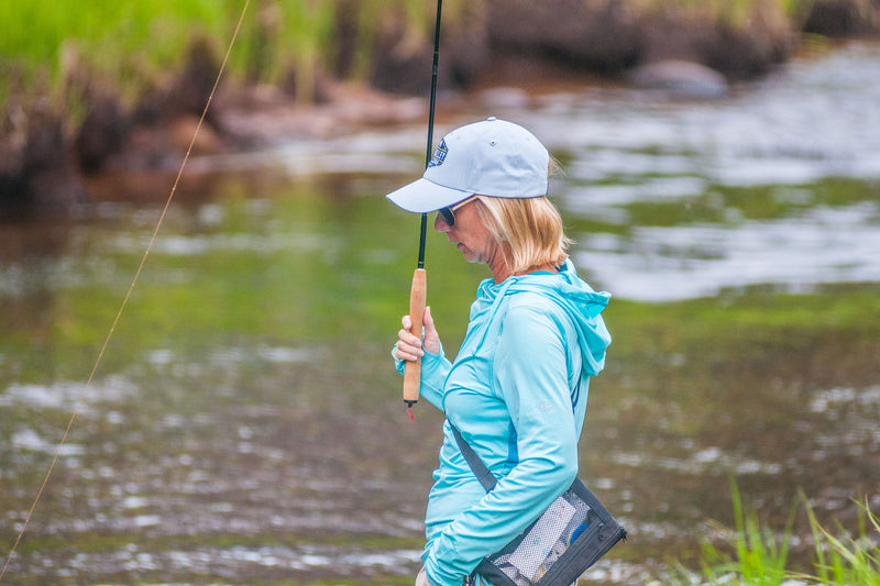Person fishing a river with a traditional Tenkara fishing rod wearing a light blue jacket and white cap.
