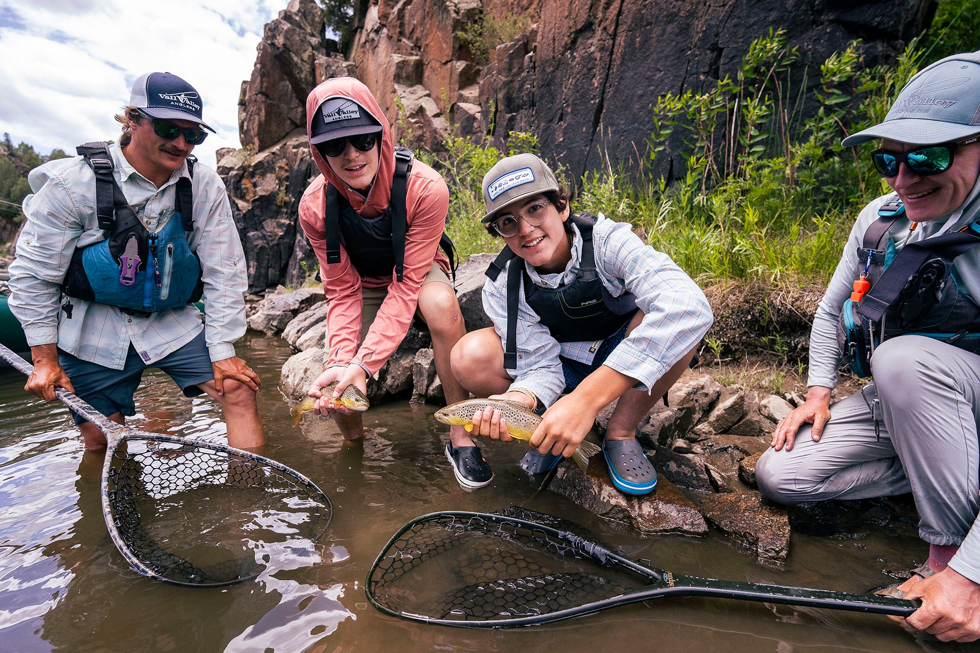 Four people in outdoor fishing gear crouch in shallow water near rocky banks, holding and netting fish. One person holds a fish while another holds a large landing net, with rock formations and greenery in the background.