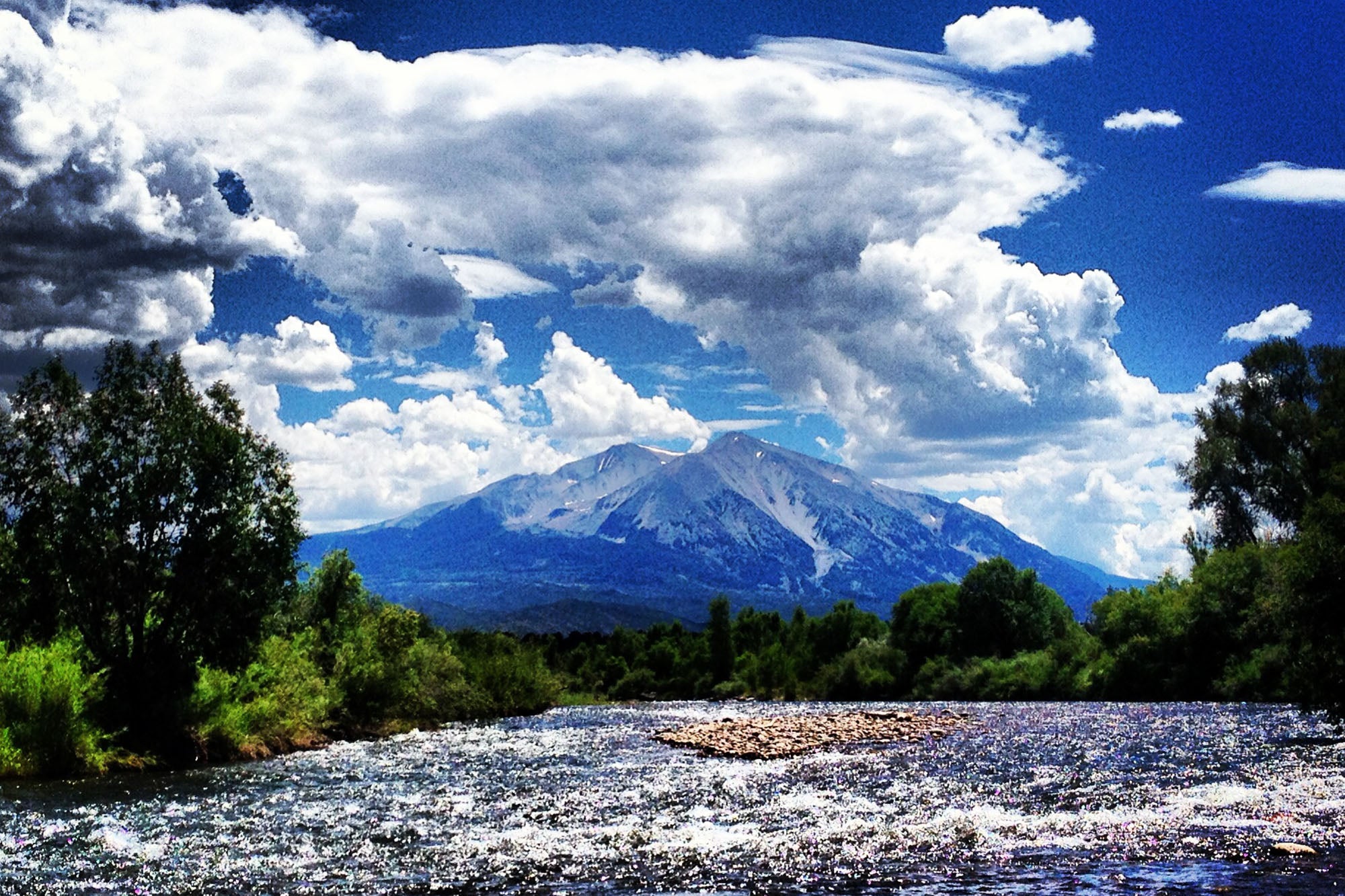Mountain range with a river and trees under a blue sky with clouds