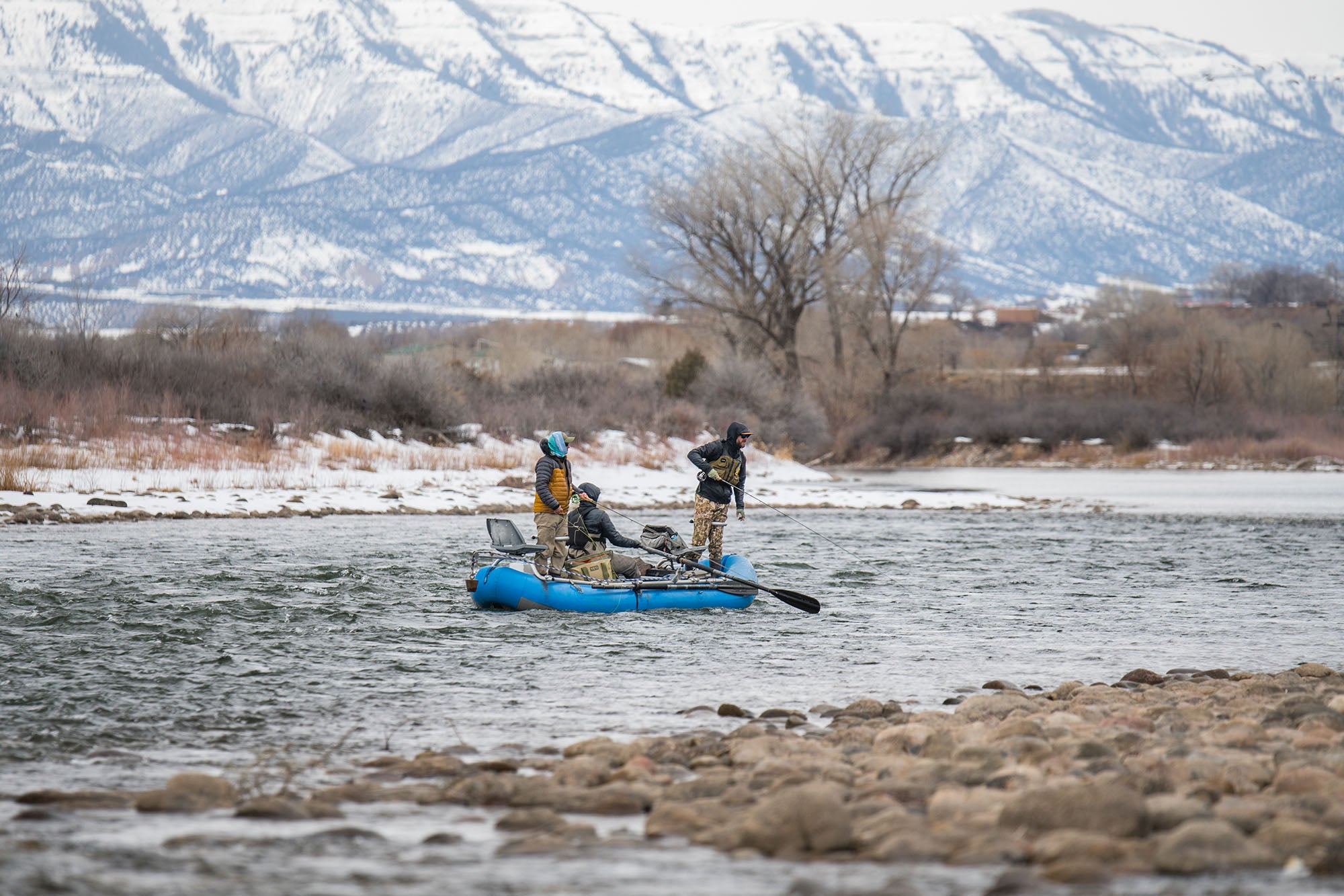Three people in a raft on the Colorado River with snow-capped mountains in the background.