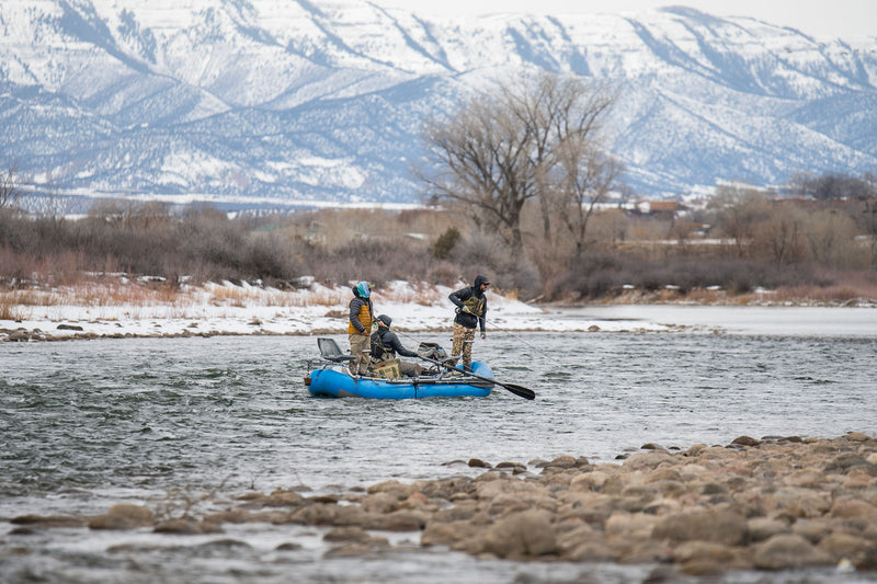 Three people in a raft on the Colorado River with snow-capped mountains in the background.